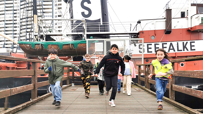 Kinder rennen auf dem Steg in Richtung Kamera. Im Hintergrund ist das Schiff Seefalke zu sehen.
