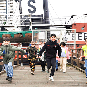 Kinder rennen auf dem Steg in Richtung Kamera. Im Hintergrund ist das Schiff Seefalke zu sehen.