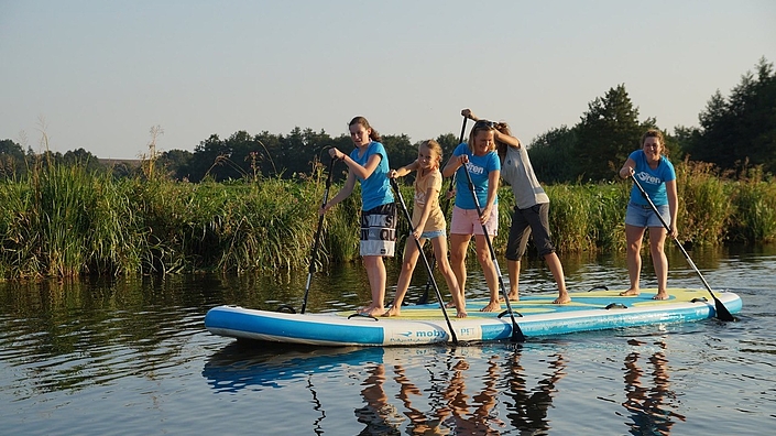 Junge Leute stehen auf einem Stand-Up_Paddle-Board und paddeln auf einem Fluss.