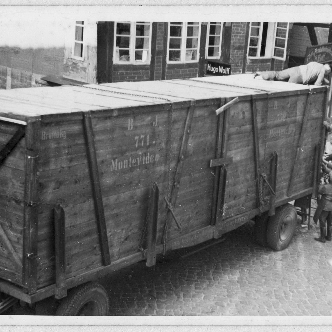 Zu sehen ist ein schwarz-weiß-Foto mit einem großen hölzernen Container auf einem Lastwagen-Anhänger. Er steht abgestellt auf einer Kopfsteinpflaster-Straße vor Fachwerkhäusern. Mehrere Männer stehen an, ein Mann liegt auf dem Container und hantieren scheinbar am Verschluss. Auf der Seite des Containers ist „771 Montevideo“ aufgedruckt.