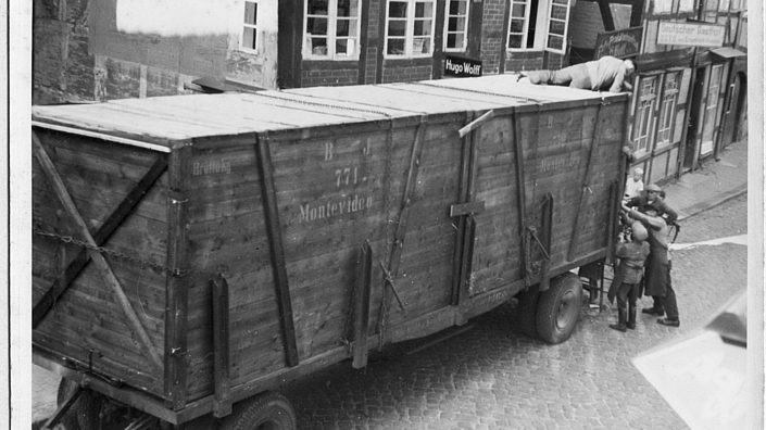 Zu sehen ist ein schwarz-weiß-Foto mit einem großen hölzernen Container auf einem Lastwagen-Anhänger. Er steht abgestellt auf einer Kopfsteinpflaster-Straße vor Fachwerkhäusern. Mehrere Männer stehen an, ein Mann liegt auf dem Container und hantieren scheinbar am Verschluss. Auf der Seite des Containers ist „771 Montevideo“ aufgedruckt.